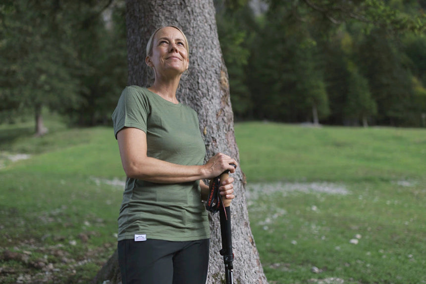 Frau im olivgrünen Merino Shirt mit Wanderstock an einen Baum gelehnt